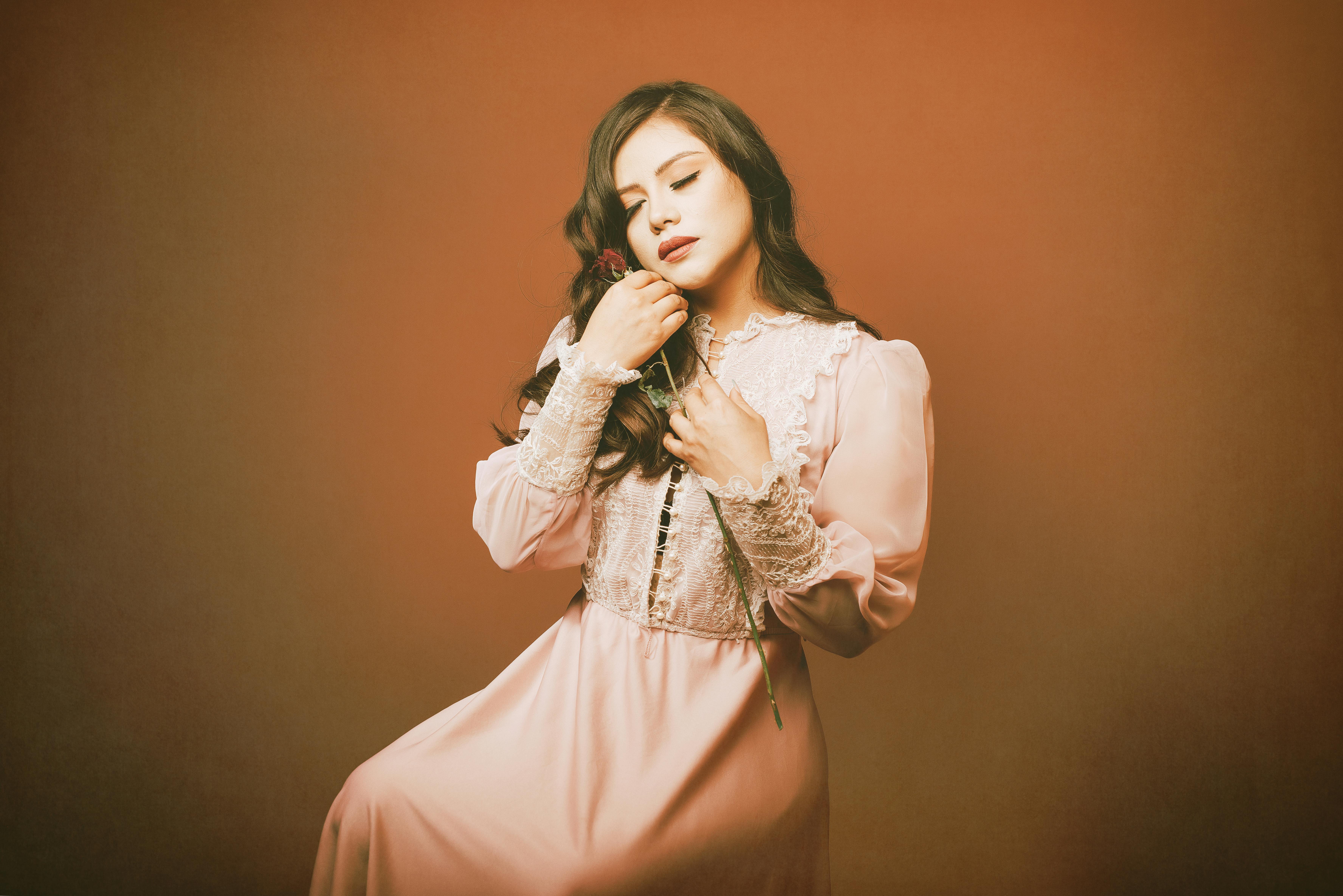 A serene woman in a vintage pink dress holds a rose, captured in a warm studio setting.